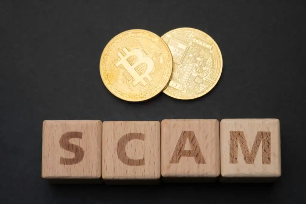 Two gold Bitcoin coins are placed above wooden blocks that spell out the word SCAM on a dark background, suggesting a warning about cryptocurrency scams.
