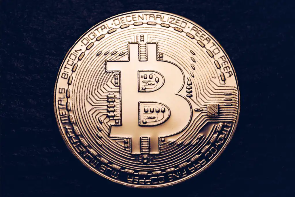 A close-up photo of a gold-colored physical Bitcoin coin with the Bitcoin symbol (₿) and circuit-like patterns, set against a dark textured background.