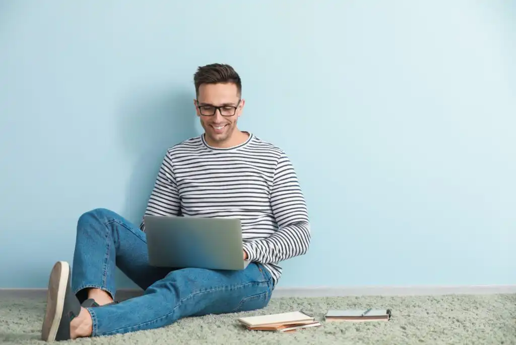 Smiling man in glasses, a striped shirt, and jeans sits on the floor against a light blue wall, using a laptop. Notebooks and a pen are on the carpet beside him.