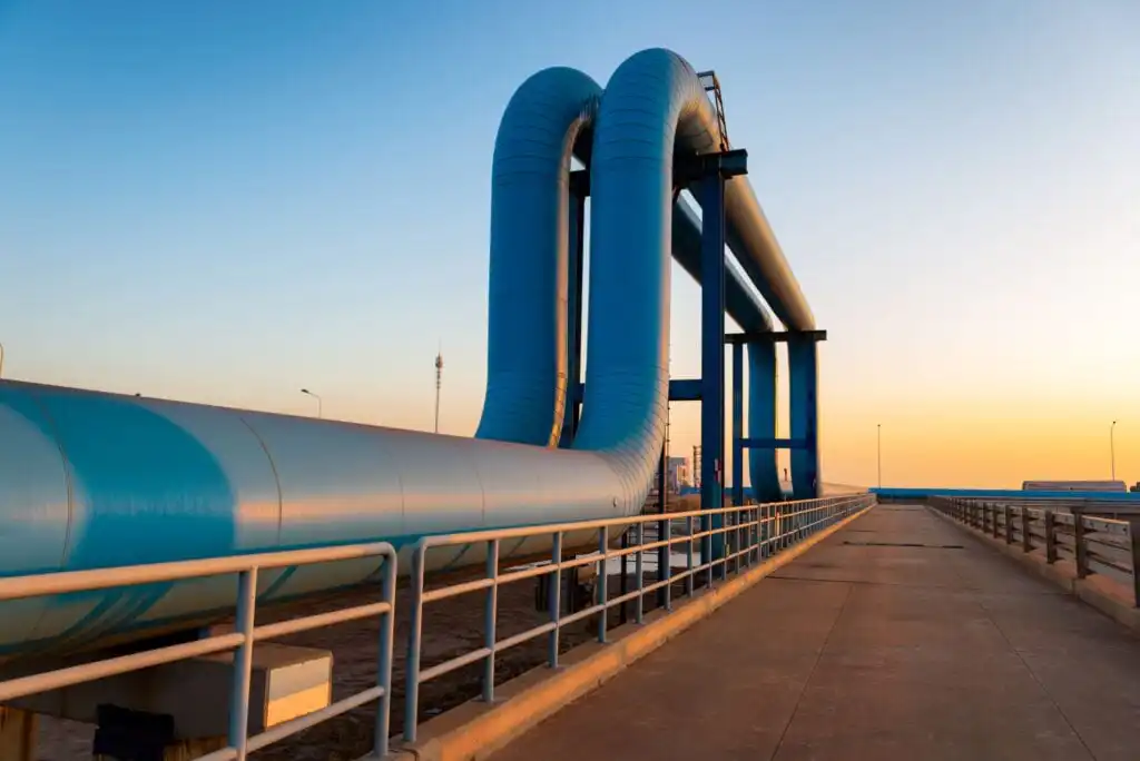 Large blue pipelines curve over a walkway at an industrial site, with metal railings on both sides and a clear sky at sunset in the background.