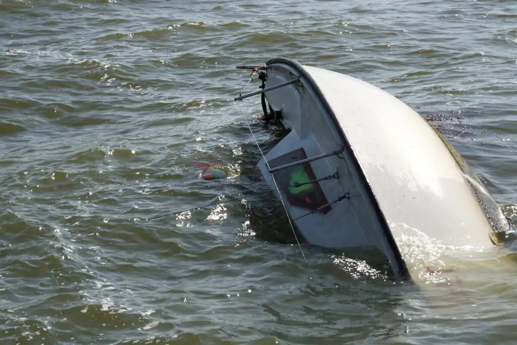 A small sailboat is capsized and partially submerged in choppy water, with only the hull and part of the sail visible above the surface.