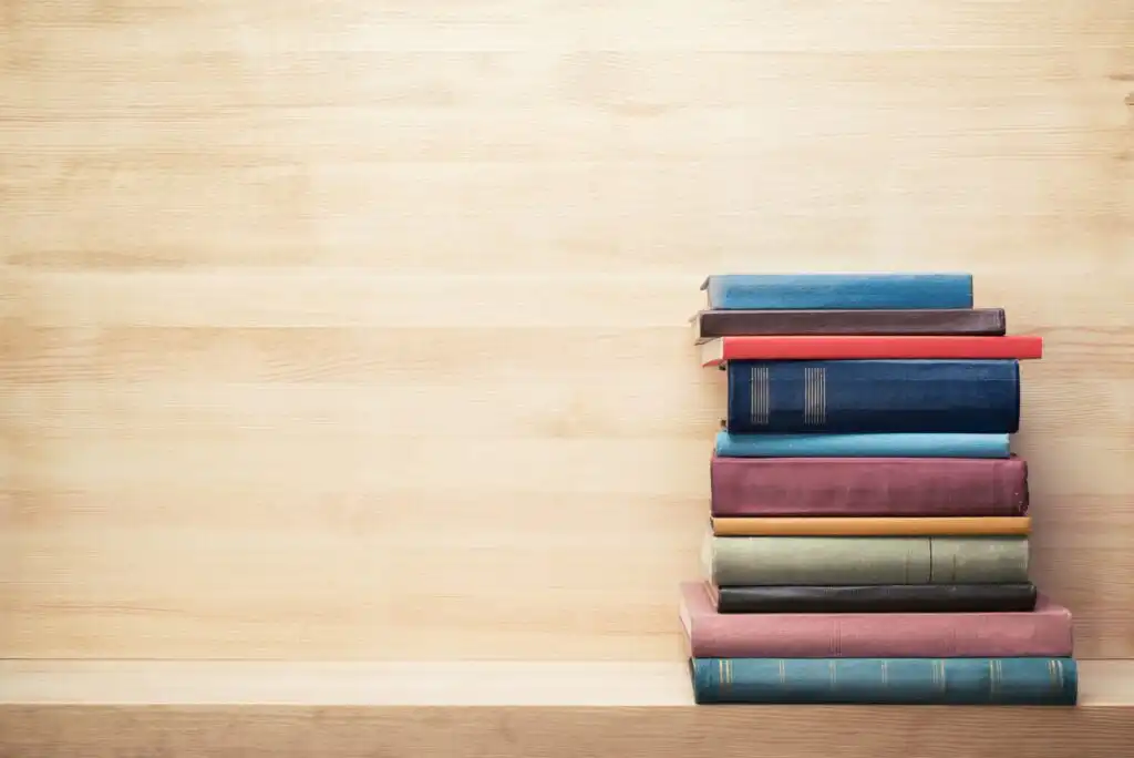 A stack of colorful hardcover books sits on a wooden surface against a light wooden background, with empty space to the left of the books.