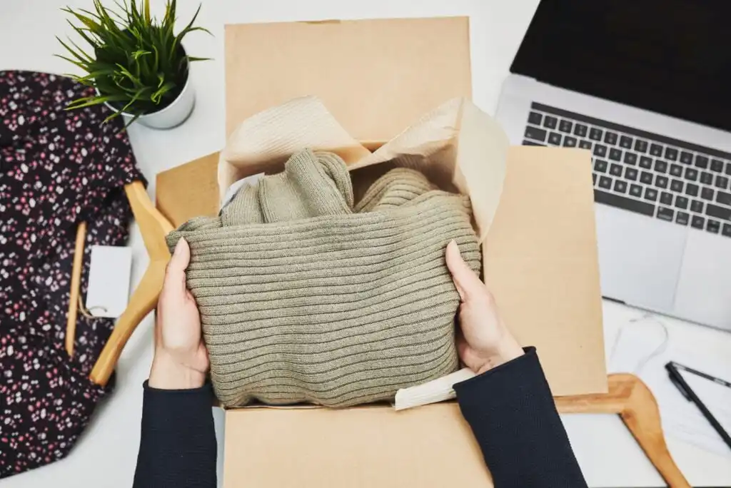 A person holds a folded, ribbed, olive-green sweater over an open cardboard box on a desk, with a patterned shirt, a laptop, a plant, and wooden hangers nearby.