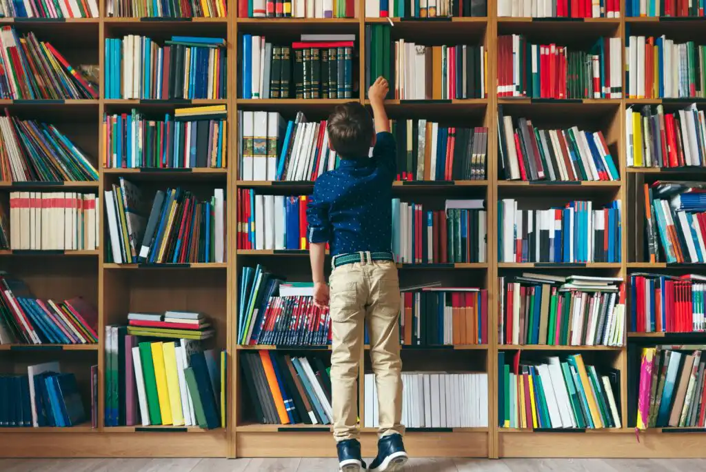 A young boy standing in front of a large bookshelf filled with books, reaching up to take a book from a high shelf.