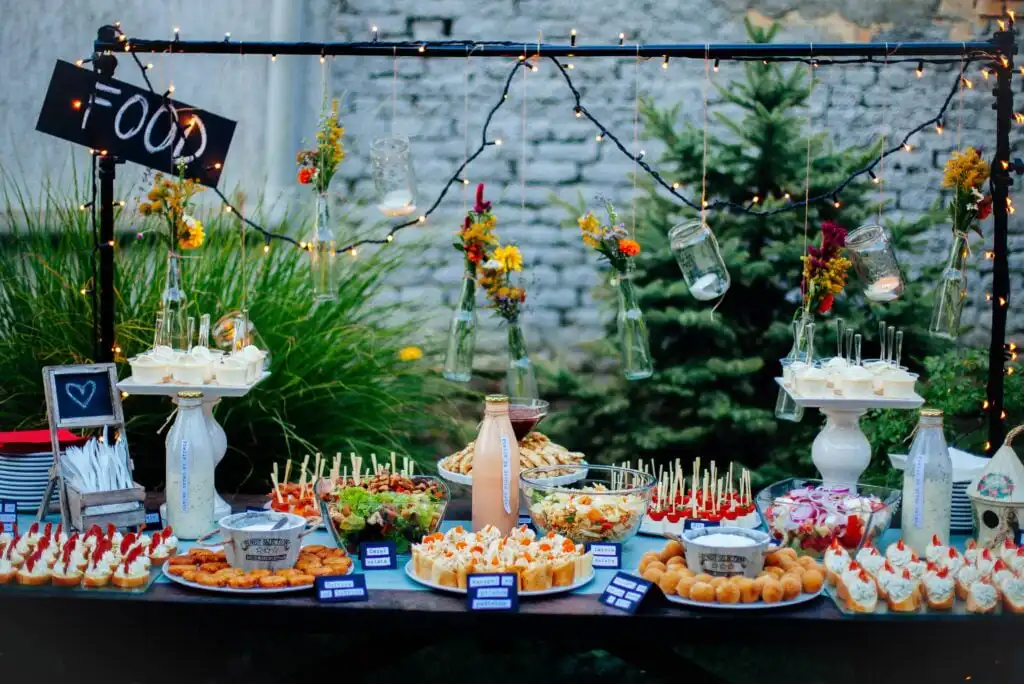 A decorated outdoor food table with various appetizers, snacks, and drinks. Flowers and jars hang above, and string lights create a festive atmosphere. Food labels and chalkboard signs are displayed on the table.