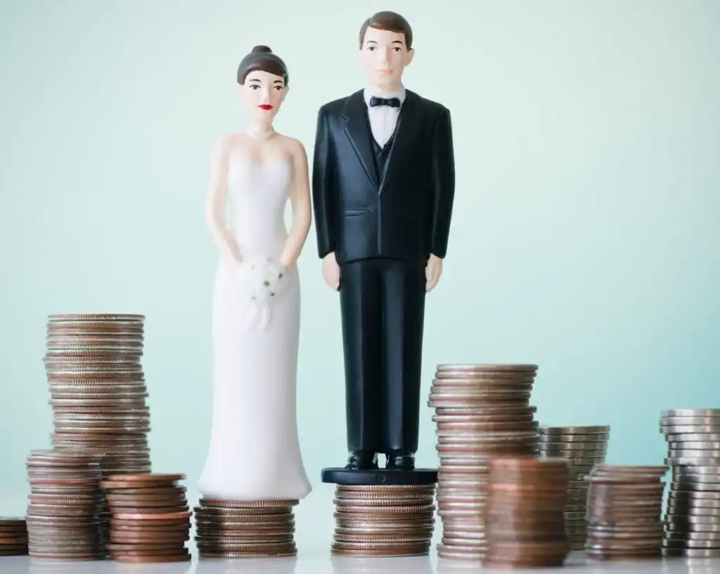 Plastic bride and groom figurines stand close together between stacks of assorted coins, symbolizing financial aspects of marriage. The background is softly lit in pale green tones.