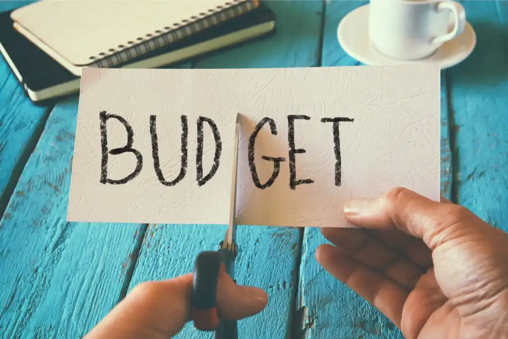A person uses scissors to cut a piece of paper with the word BUDGET written on it, symbolizing budget cuts. A notebook, pencil, and coffee cup sit on a turquoise wooden table in the background.