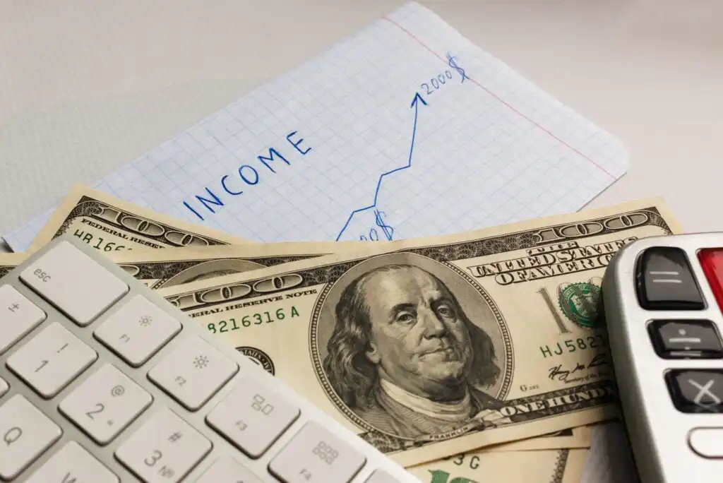 A close-up of US hundred-dollar bills, a white keyboard, and a calculator, with a graph labeled “Income” showing an upward trend on graph paper in the background.