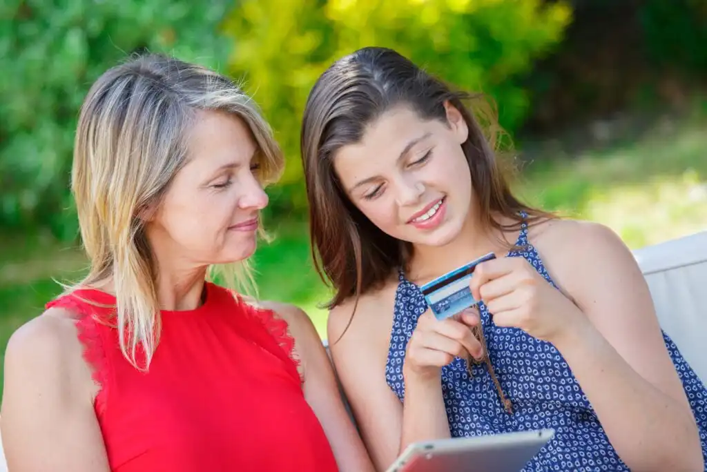 A woman in a red sleeveless top sits next to a girl in a blue dress. The girl is holding a credit card and using it with a tablet as they both smile outdoors on a sunny day.