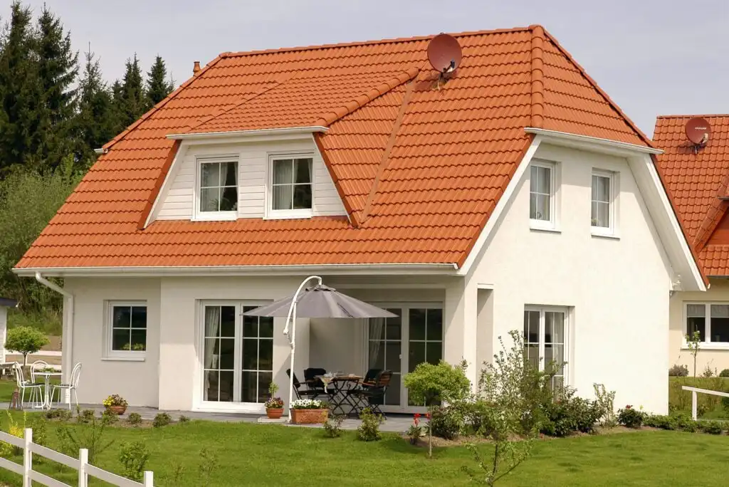 A modern white house with a red tiled roof, dormer windows, and a satellite dish. There is a patio with outdoor furniture and a gray umbrella, surrounded by a well-kept lawn and garden.