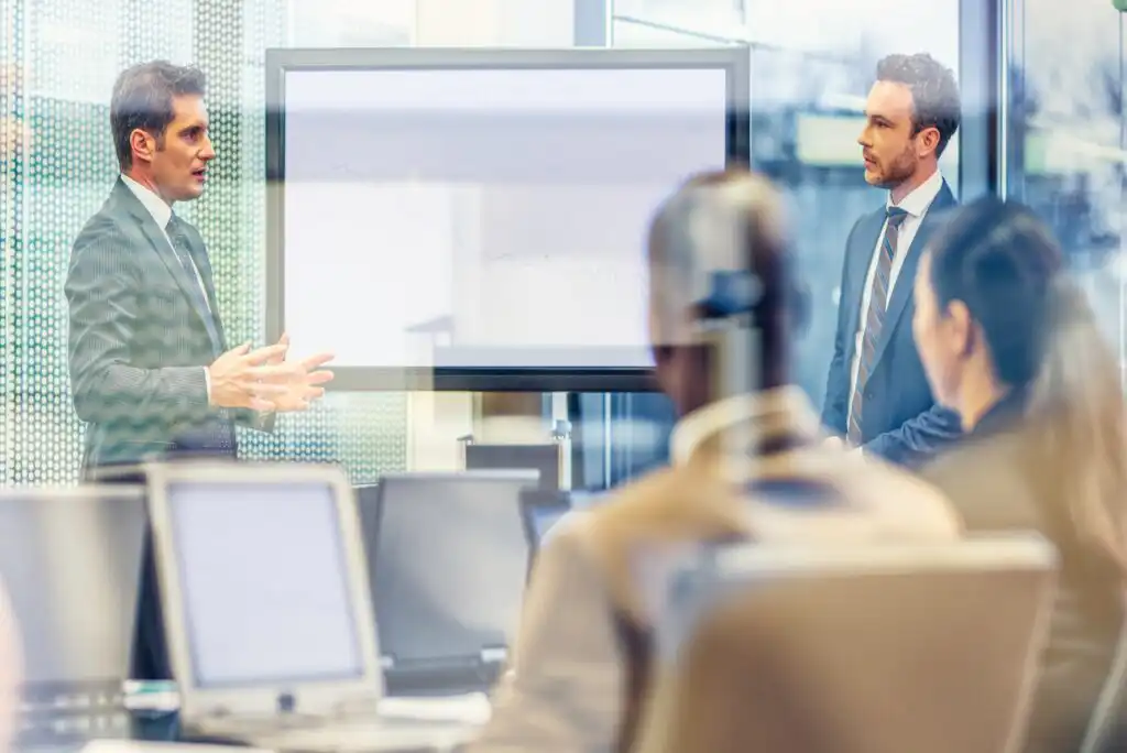 Two men in business suits lead a meeting in a modern conference room, standing by a screen with a faint graph. Several colleagues sit at a table with laptops, listening attentively. The scene is viewed through glass.