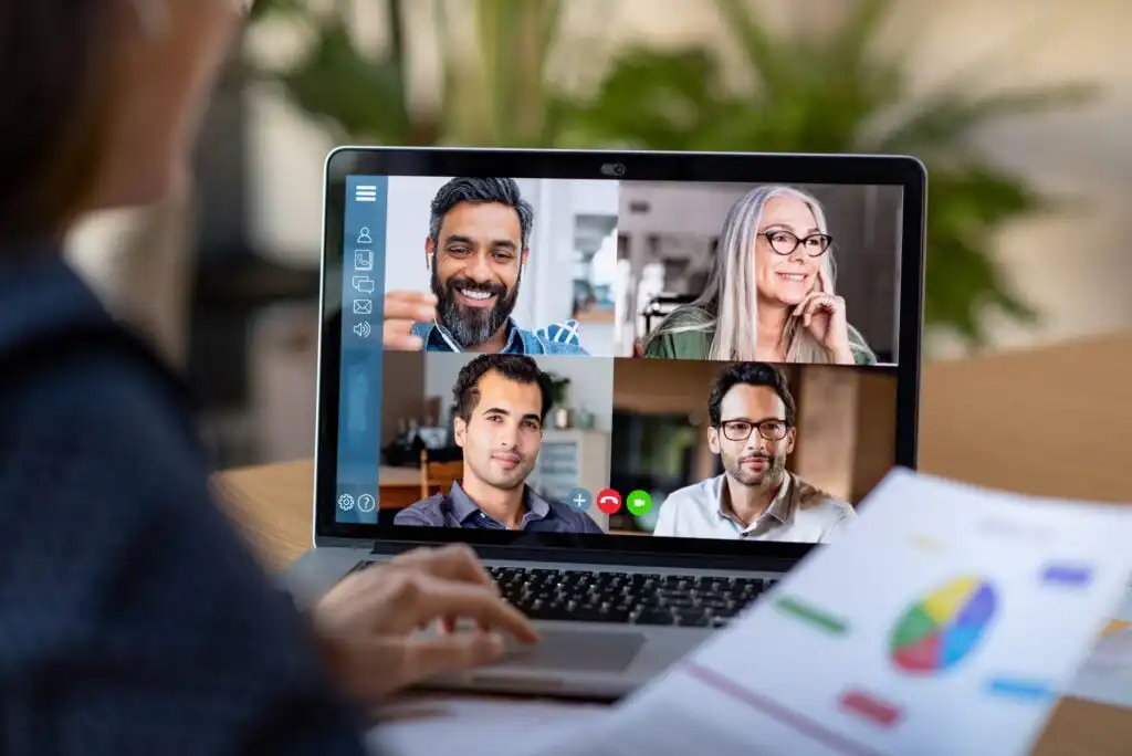 A person participates in a video conference on a laptop with four people visible on the screen. The person holds a document with a colorful pie chart. The background is blurred, focusing on the meeting.