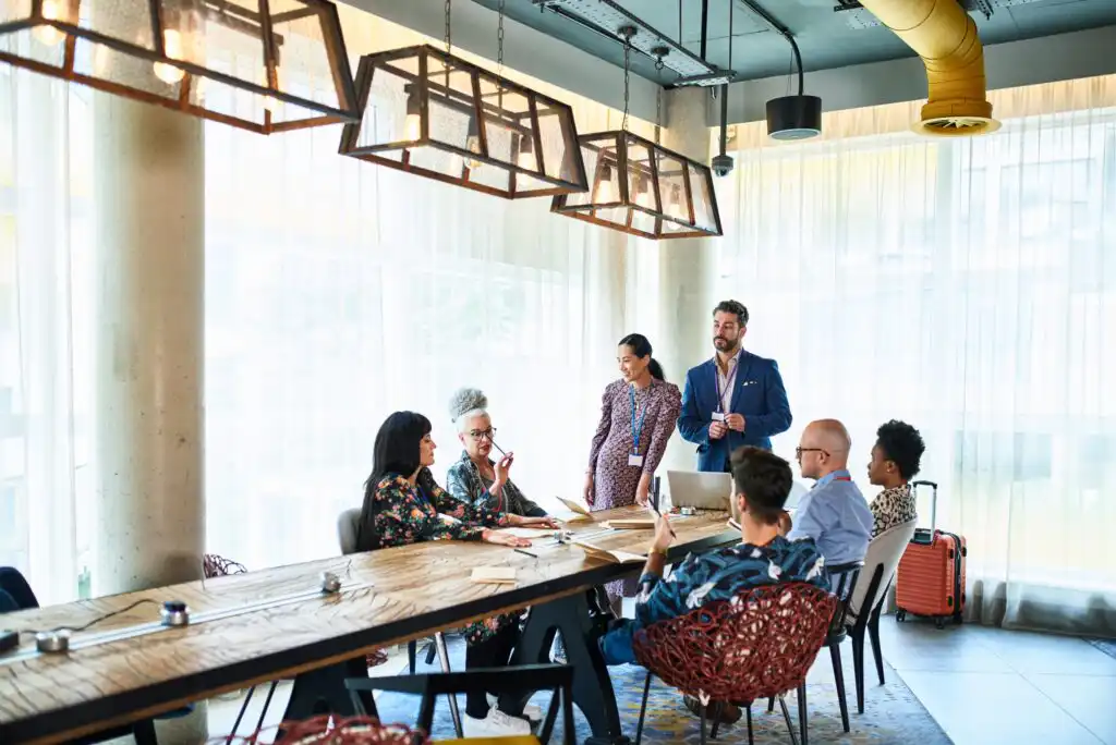 Seven people are gathered in a modern conference room with large windows, sitting and standing around a long table, engaged in discussion. Laptops, papers, and coffee cups are on the table.