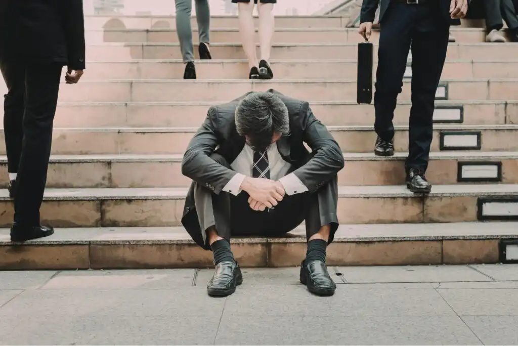 A man in a suit sits on outdoor steps with his head down and arms resting on his knees, looking dejected, while people in business attire walk past him.