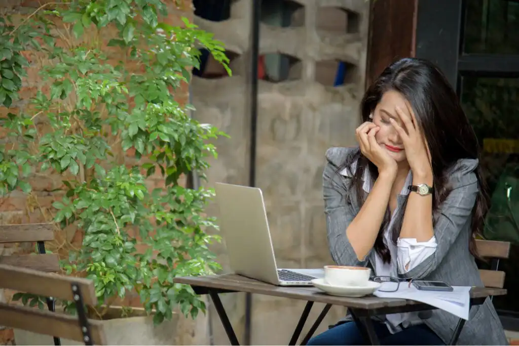 A woman sits at an outdoor table with a laptop, cup of coffee, and phone, resting her face in her hand and appearing stressed or tired. Lush green plants and a brick wall are in the background.