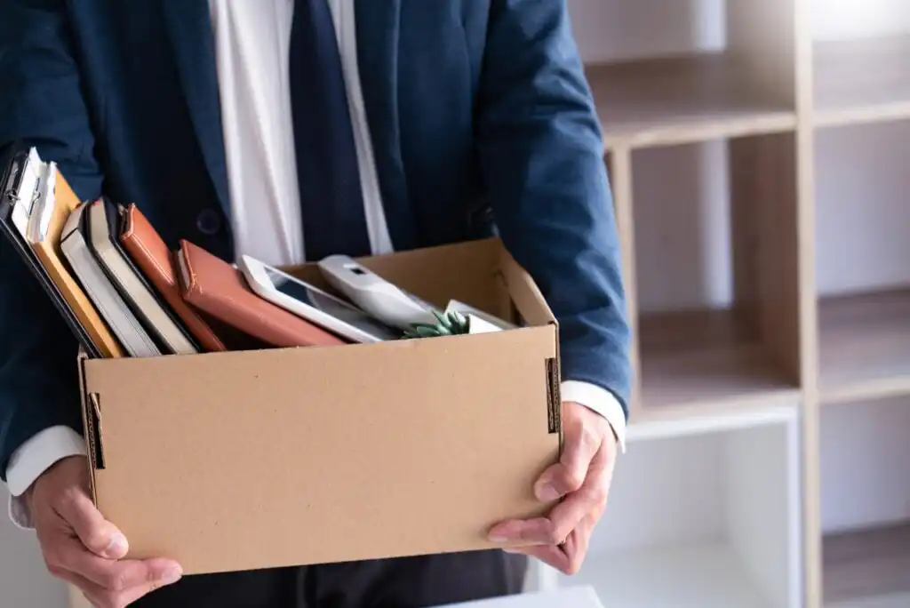 A person in a suit holds a cardboard box filled with office supplies, including notebooks, a phone, and a potted plant, suggesting they are clearing out their workspace or leaving their job.