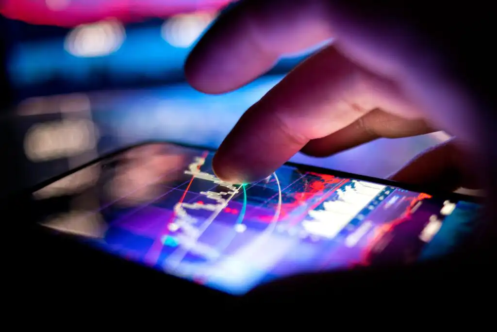 Close-up of a hand touching a smartphone screen displaying colorful financial graphs and charts, indicating stock market or trading activity in a dark environment.