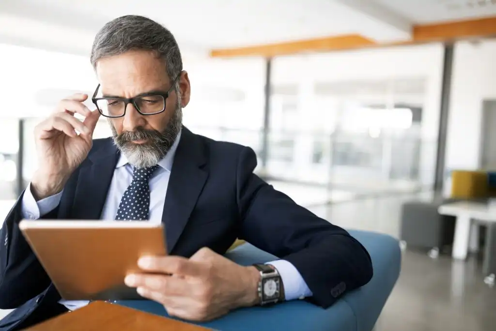 A middle-aged man in a suit and tie sits indoors, adjusting his glasses while reading from a tablet. He has gray hair, a beard, and appears focused, with a modern office setting in the background.