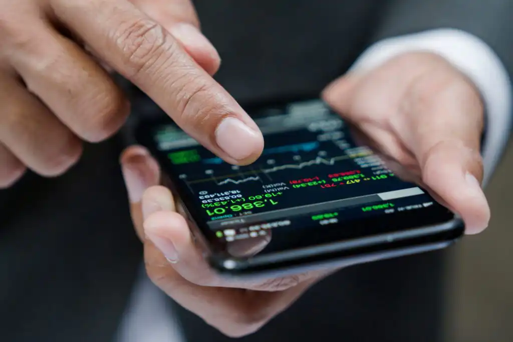 A person in a suit holds a smartphone displaying a stock market app with graphs and numbers, using their index finger to interact with the screen.