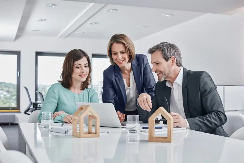 Three business professionals sit at a table with a laptop, documents, and small wooden house models, discussing work in a modern, bright office setting.