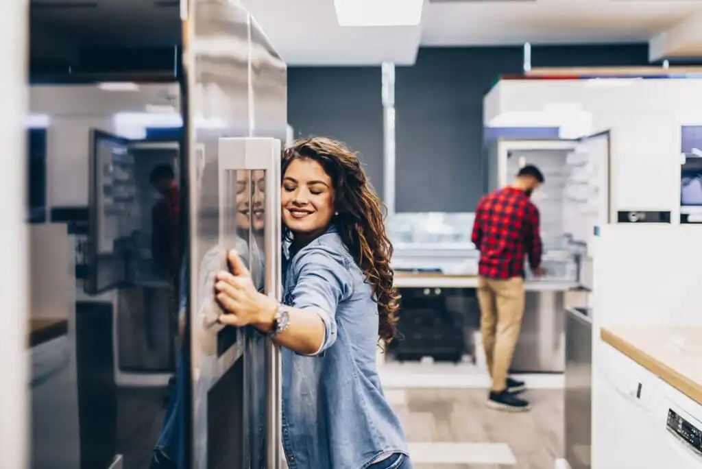 A smiling woman hugs a stainless steel refrigerator in an appliance store, while a man in a red plaid shirt examines another refrigerator in the background.