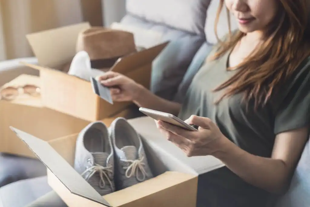A woman sits on a couch holding a smartphone and a credit card, with an open box containing new gray sneakers on her lap and another box beside her.