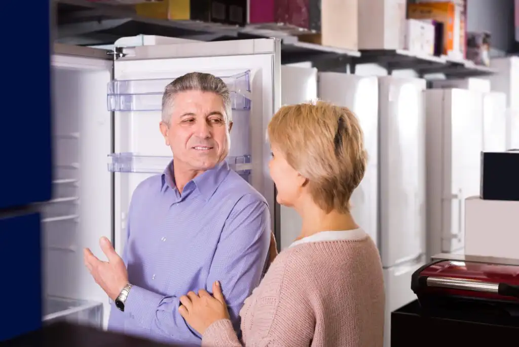 An older man and woman stand together in an appliance store, smiling as they look at an open refrigerator, appearing to discuss or consider purchasing it. Other refrigerators are visible in the background.
