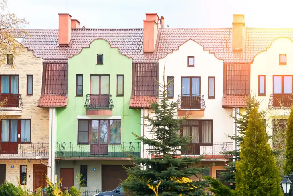 A row of colorful townhouses with distinctive gabled roofs, balconies, and large windows, surrounded by green trees and shrubs on a sunny day.