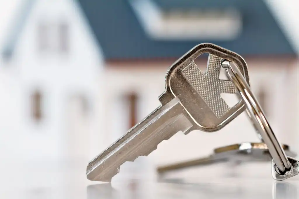 A close-up of a house key on a keyring, with a blurred image of a house in the background. The focus is on the key, symbolizing home ownership or real estate.