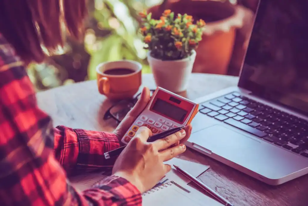 A person in a red plaid shirt uses a calculator at a desk with a laptop, notebook, pen, cup of coffee, and a small potted plant, with sunlight streaming in the background.