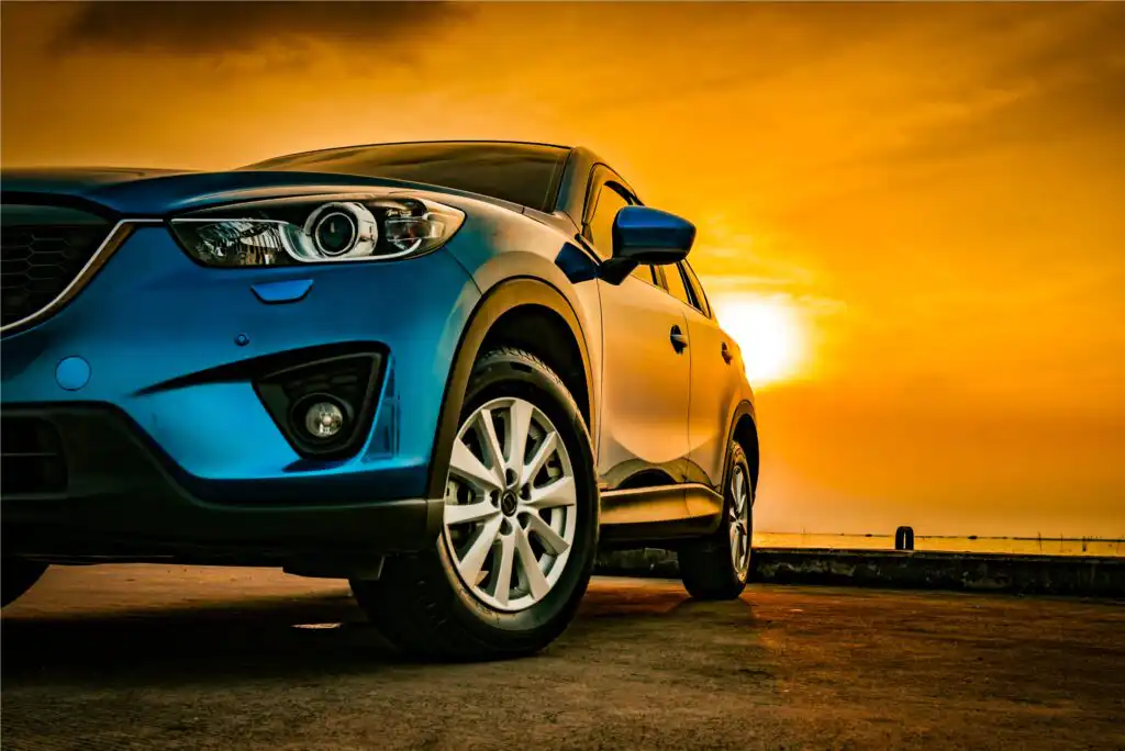 A blue SUV parked on a road at sunset, with the sun low in the orange sky, casting warm light and reflections on the car’s shiny surface.
