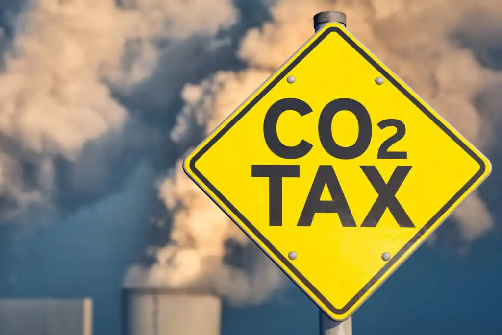 A yellow road sign with CO₂ TAX written on it stands in front of industrial chimneys emitting large clouds of smoke into the sky.