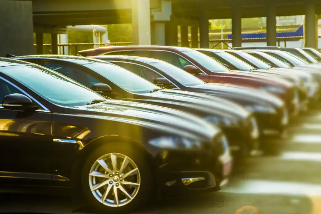 A row of parked cars in a parking lot, with sunlight shining on their hoods and windshields. The vehicles are lined up closely, and the background shows a covered parking structure.