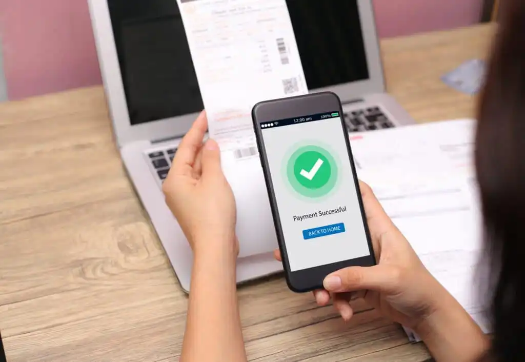 A person holds a phone displaying a “Payment Successful” message with a green check mark while holding a bill in front of an open laptop on a wooden desk.