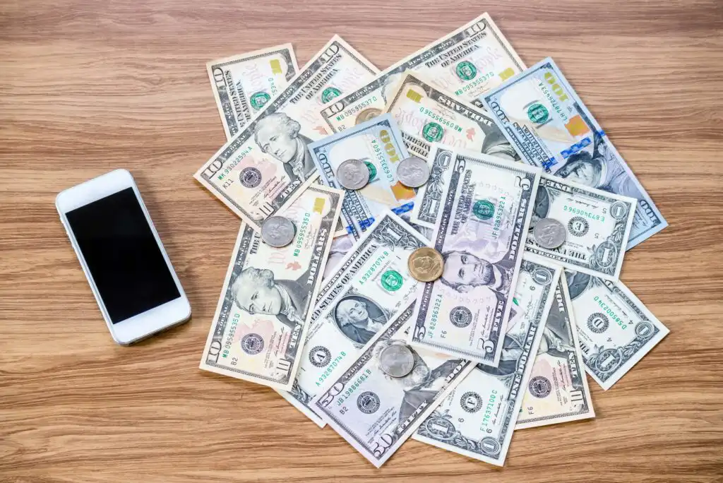 A smartphone and a scattered pile of various US dollar bills and coins are placed on a wooden surface.
