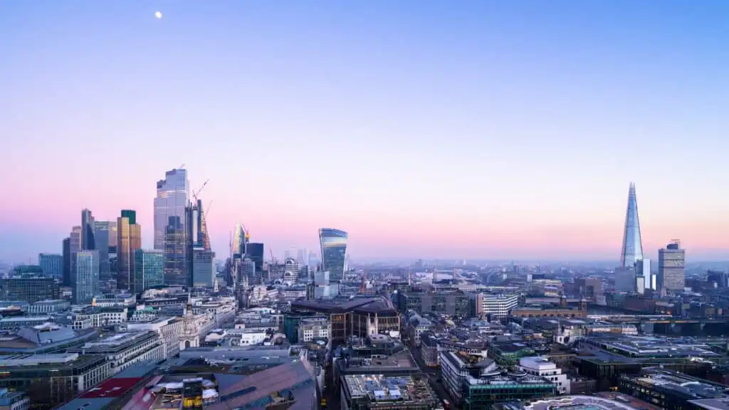 Panoramic view of London’s city skyline at dusk, featuring modern skyscrapers like the Shard and Walkie Talkie building, with a pastel sky and moon visible above the urban landscape.