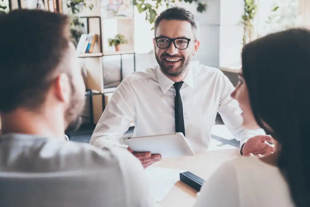A smiling man in glasses and a white shirt holds a tablet while talking to two people seated across from him in a bright, modern office with plants and shelves in the background.