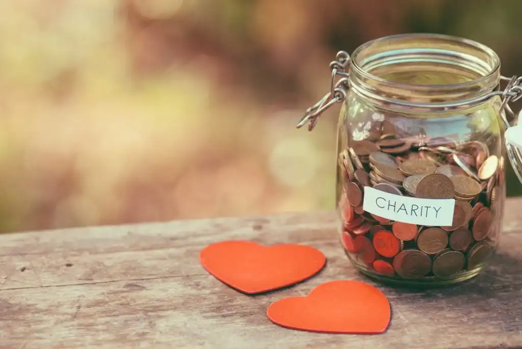 A glass jar labeled charity filled with coins sits on a wooden surface outdoors, next to two red paper hearts, symbolizing donation and generosity.