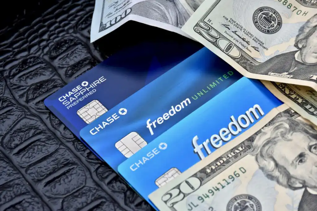 Three Chase credit cards (Sapphire Preferred, Freedom Unlimited, and Freedom) are arranged on a black textured surface next to several U.S. twenty-dollar bills.