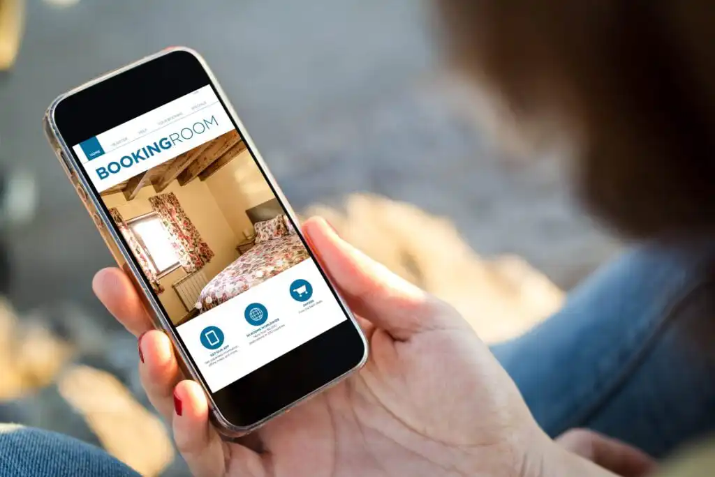 A person holds a smartphone displaying a hotel booking app, showing a bedroom with floral curtains and bedspread, under the header BOOKINGROOM. The background is blurred.