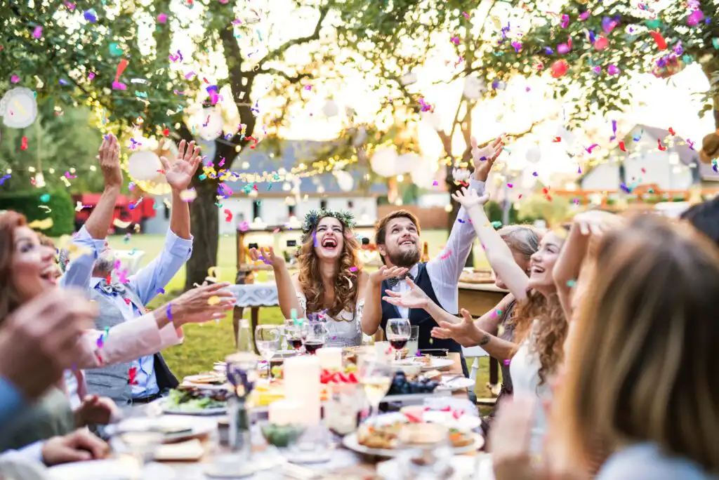 A group of people sits around an outdoor table, laughing and celebrating as colorful confetti falls. The table is set with food and drinks, and the scene is bright and festive, with trees and string lights in the background.