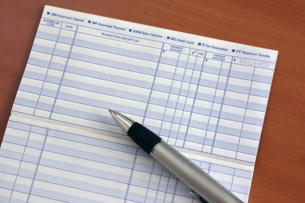 A close-up of a checkbook register with blank columns and rows for recording transactions. A silver and black pen rests diagonally on the paper, all placed on a wooden surface.