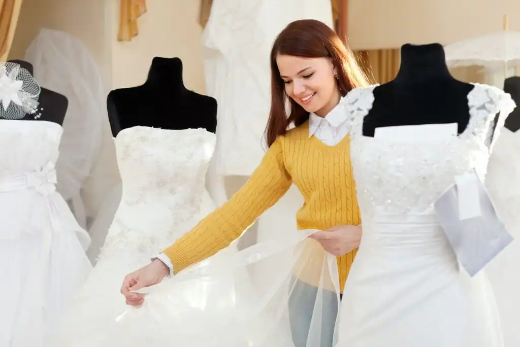 A smiling woman in a yellow sweater examines the skirt of a wedding dress on a mannequin in a bridal shop, surrounded by other white wedding dresses on display.