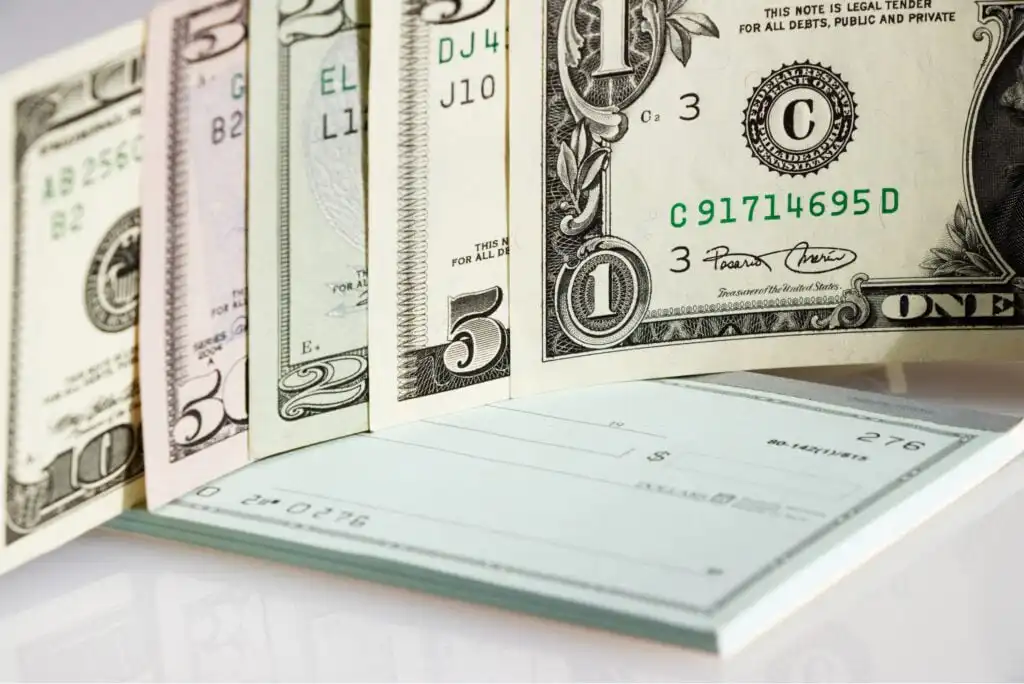 Several US dollar bills in various denominations are fanned out next to an open checkbook with blank checks, all placed on a reflective surface.