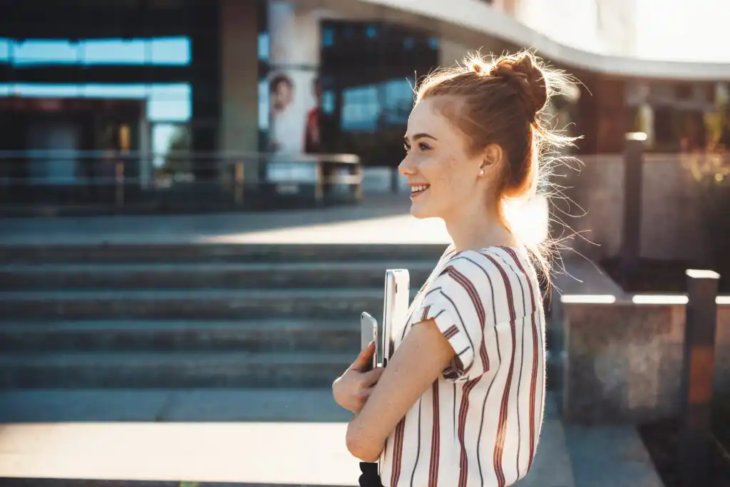 A young woman with red hair in a bun, wearing a striped shirt, stands outside holding books and smiling. She is in front of a building with steps in the background on a sunny day.