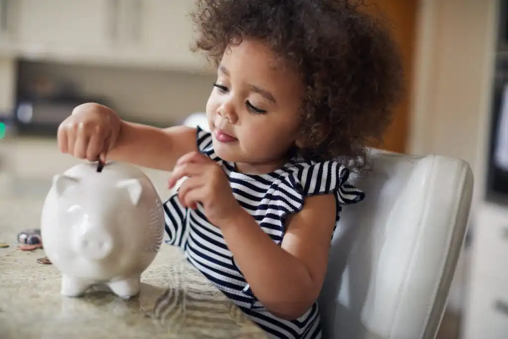A young child with curly hair wearing a striped dress is sitting at a table, smiling and placing a coin into a white piggy bank.