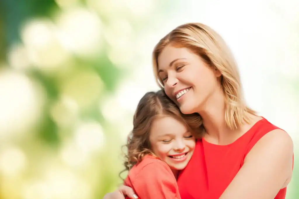 A smiling woman and a young girl, both wearing red, hug each other happily outdoors with a blurred green and white background.