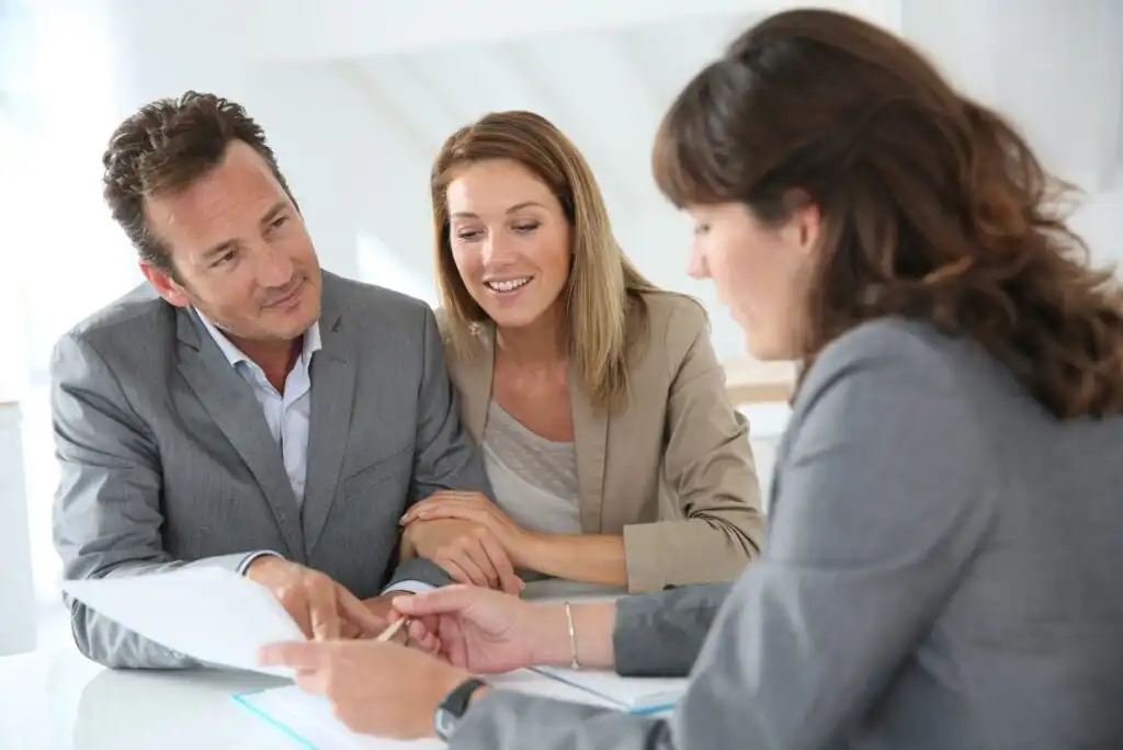 A woman in a suit is showing documents to a smiling couple seated across from her at a desk. The couple appears attentive and engaged in the discussion. All three are dressed in business attire.