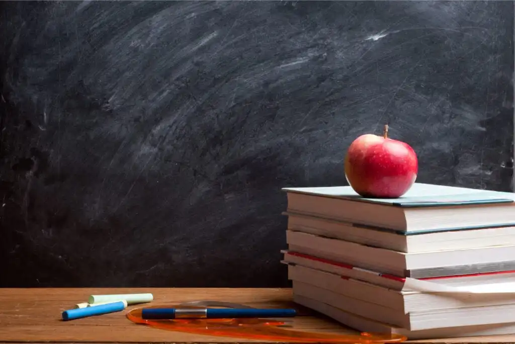 A stack of books with a red apple on top sits on a wooden desk in front of a blank chalkboard, alongside some colored chalk and a pair of orange glasses.