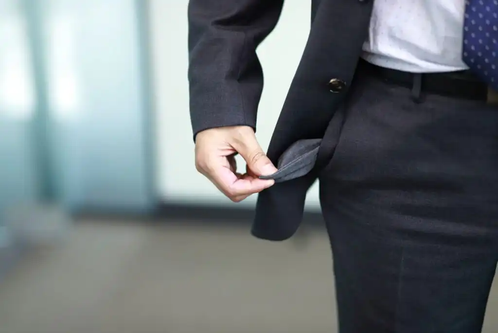 A person in a business suit is pulling out their empty pants pocket, suggesting they have no money. The background is out of focus.